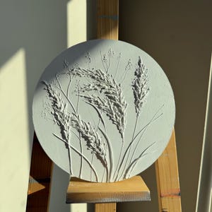 May include: A circular, off-white relief sculpture showing detailed tall grasses and seed heads. The artwork is displayed on a wooden easel. Sunlight casts shadows across the textured surface, emphasising the botanical forms.