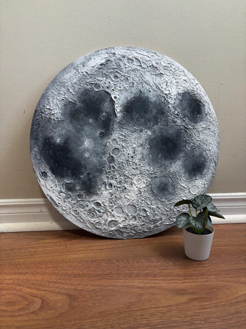 Peut inclure: Une grande peinture ronde de la lune, de couleur blanche et grise, avec une surface textur&eacute;e. La peinture est sur un sol en bois avec une petite plante en pot au premier plan.