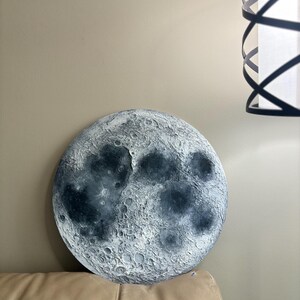 Peut inclure: Une d&eacute;coration murale ronde en forme de lune, de couleur gris et blanc, avec une surface textur&eacute;e. La lune est accroch&eacute;e &agrave; un mur au-dessus d'un canap&eacute; beige.