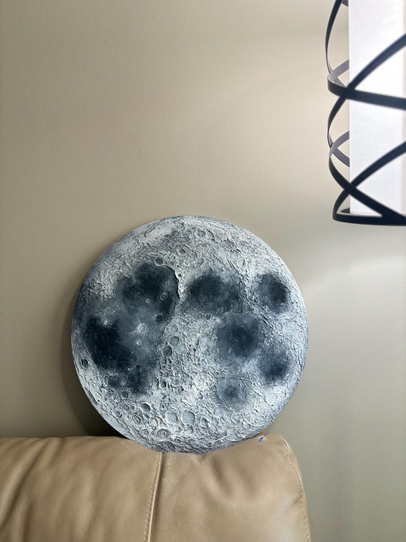 Peut inclure: Une d&eacute;coration murale ronde en forme de lune, de couleur gris et blanc, avec une surface textur&eacute;e. La lune est accroch&eacute;e &agrave; un mur au-dessus d'un canap&eacute; beige.