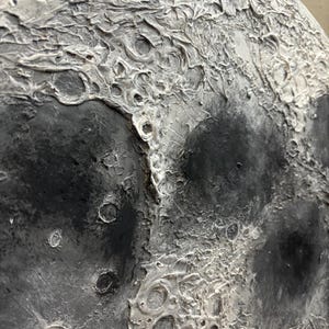Peut inclure: Gros plan sur une peinture de lune textur&eacute;e, grise et blanche. La surface de la lune est peinte avec une vari&eacute;t&eacute; de textures, y compris des crat&egrave;res et des cr&ecirc;tes.