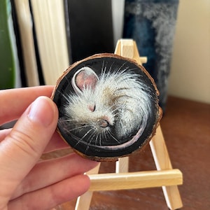 May include: A small, white mouse painted on a round wooden slice. The mouse is curled up and sleeping. The background is black.