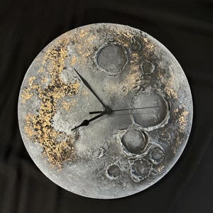 Peut inclure: Une horloge en forme de lune grise et blanche avec des accents dorés et des aiguilles noires. Le cadran de l'horloge est texturé pour ressembler à la surface de la lune.