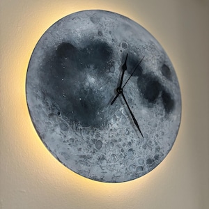 May include: A round, gray and white moon-shaped clock with black hands on a white wall. The clock has a light halo around it.