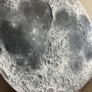 Peut inclure: Gros plan d'une sculpture ronde de lune textur&eacute;e en gris et blanc. La surface de la lune est d&eacute;taill&eacute;e avec des crat&egrave;res et d'autres caract&eacute;ristiques lunaires.