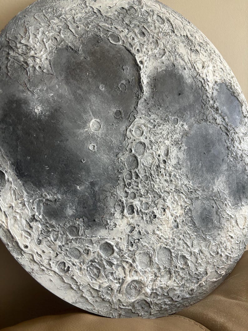 Peut inclure: Gros plan d'une sculpture ronde de lune textur&eacute;e en gris et blanc. La surface de la lune est d&eacute;taill&eacute;e avec des crat&egrave;res et d'autres caract&eacute;ristiques lunaires.