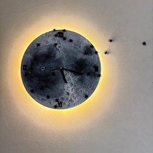 Può includere: Un orologio da parete rotondo con un design a forma di luna. Il quadrante mostra una rappresentazione dettagliata della superficie lunare, con numeri romani e lancette nere. L'orologio è illuminato da un bagliore giallo caldo sul bordo.