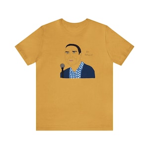 Puede incluir: Camiseta de color amarillo mostaza con una ilustración de dibujos animados de un hombre sosteniendo un micrófono. El hombre lleva una blazer azul sobre una camisa de lunares blancos y azules. El texto "by GOLLY!" está impreso en la camiseta.