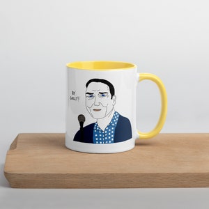 May include: A white ceramic mug with a yellow rim and handle. The mug features a cartoon illustration of a man with a microphone, saying "By Golly!"