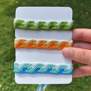 以下が含まれることがあります： 緑、オレンジ、青色の波模様の3つの手作りの友情ブレスレット。各ブレスレットは白いカードに表示されています。ブレスレットは織り糸で作られており、航海のテーマを持っています。