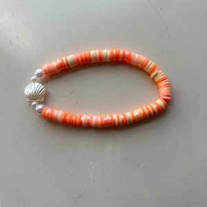 May include: A beaded bracelet featuring orange, peach, and cream-colored disc beads. The bracelet is accented with a silver seashell charm and small white pearls. The bracelet is approximately 7 inches in length.