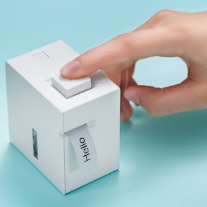 May include: A white cube-shaped device with a button on top and a slot on the side. A label with the word "Hello" is emerging from the slot. The device is on a light blue surface.