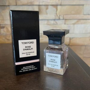May include: A black rectangular box and a clear perfume bottle with a black cap. Both feature a white label with the text "TOM FORD ROSE D'AMALFI EAU DE PARFUM 30 ML". The bottle is 1 fl oz.