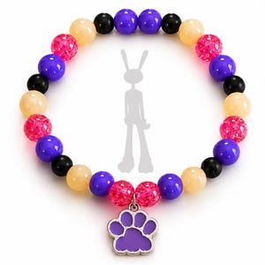 Puede incluir: Una pulsera de cuentas con un patrón repetitivo de cuentas redondas en morado, negro, amarillo claro y rosa intenso texturizado. Un dije de metal plateado con forma de huella y centro de esmalte morado cuelga de la parte inferior de la pulsera.