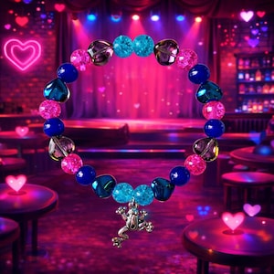 Puede incluir: Una pulsera de cuentas con un dije de rana plateada. Presenta cuentas azules, rosas y transparentes. El fondo es una escena de escenario borrosa con iluminación rosa y azul.