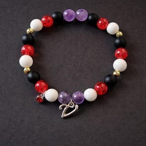 May include: A beaded bracelet featuring a mix of red, white, black, and purple beads. The bracelet includes a silver charm with a small red jewel and a silver pendant. The beads are interspersed with gold-colored spacers.
