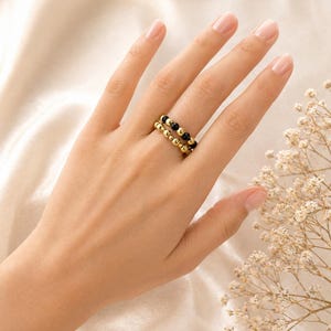 Puede incluir: Dos anillos dorados con cuentas, uno con cuentas negras, se llevan en un dedo. Los anillos se colocan sobre un fondo suave blanquecino con una ramita de flores secas en la esquina. Los anillos son de diseño sencillo y elegante.