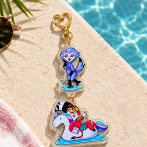 May include: A double-layered acrylic keychain featuring two cartoon characters. The top charm shows a blue owl-like figure standing in water. The bottom charm depicts a red and white character riding a white unicorn pool float with blue accents.