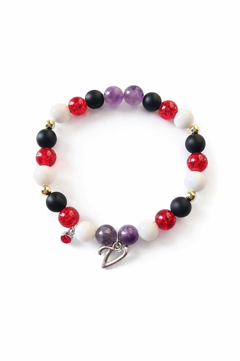 May include: A beaded bracelet featuring a mix of red, white, black, and purple beads. The bracelet includes a silver heart charm and a small red charm. The beads are interspersed with gold-colored spacers.