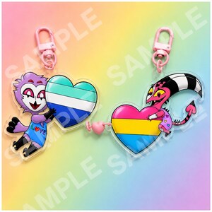 May include: Two acrylic keychains connected by a small pink heart charm. The left keychain features a purple character holding a heart with teal, white, and blue stripes. The right keychain shows a pink character holding a heart with pink, yellow, and blue stripes.