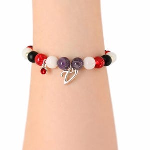 May include: A beaded bracelet featuring a mix of red, white, black, and purple beads. The bracelet has a silver charm in the shape of a V and a small red charm. The bracelet is worn on a wrist.