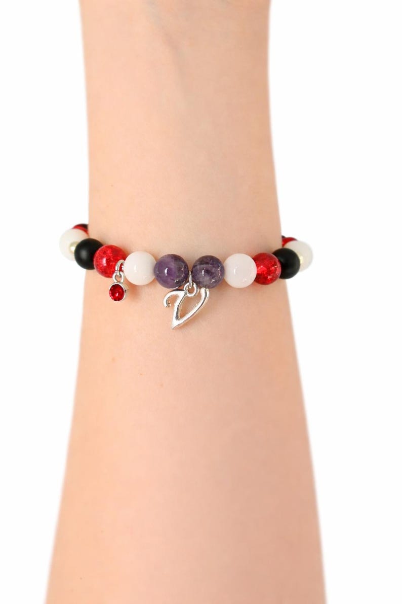 May include: A beaded bracelet featuring a mix of red, white, black, and purple beads. The bracelet has a silver charm in the shape of a V and a small red charm. The bracelet is worn on a wrist.