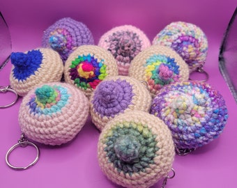 May include: A collection of crocheted nipple keychains in various colors, including pink, purple, blue, and multicolored yarn. Each keychain features a detailed nipple design and a silver-colored keyring. The keychains are arranged on a solid purple background.