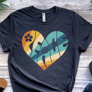 May include: A dark grey t-shirt with a retro-style heart design featuring the word "PEACE" in a colorful, layered font. The heart is surrounded by flowers and stripes.