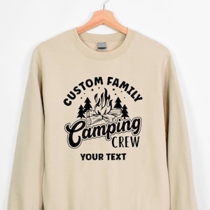 Pode incluir: Uma camisa de moletom bege com um design gr&aacute;fico preto que diz "Custom Family Camping Crew" com uma fogueira e &aacute;rvores. O texto "Your Text" est&aacute; abaixo do gr&aacute;fico.