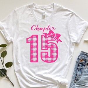 May include: White t-shirt with the text "Chapter 15" in pink script and a pink bow. The number 15 is in a pink and white plaid pattern. The shirt is styled with denim shorts and a starfish.