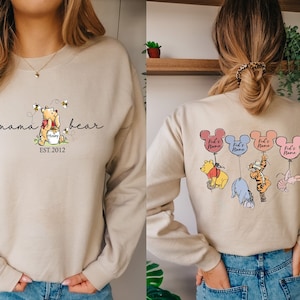 May include: Two beige sweatshirts with Winnie the Pooh themed designs. The front of the sweatshirt has a Winnie the Pooh illustration with the text 'mama bear EST. 2012'. The back of the sweatshirt has four Winnie the Pooh characters holding balloons with the text 'Kid's Name' on each balloon.