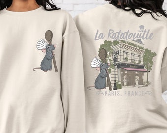 May include: A beige sweatshirt with a cartoon rat chef holding a wooden spoon on the front and a graphic of a Parisian restaurant with the text "La Ratatouille, Paris, France" on the back.
