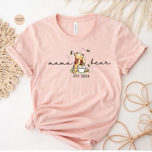 Pu&ograve; includere: T-shirt color pesca chiaro con un disegno di Winnie the Pooh, le parole "mama bear" e "EST. 2024". Il disegno include api e un vasetto di miele. La maglietta &egrave; in materiale morbido.