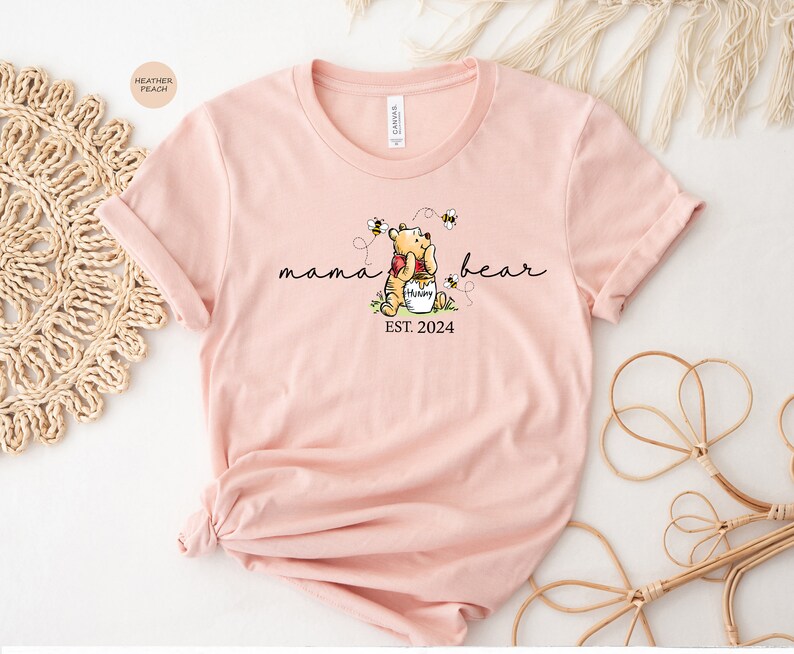 Pu&ograve; includere: T-shirt color pesca chiaro con un disegno di Winnie the Pooh, le parole "mama bear" e "EST. 2024". Il disegno include api e un vasetto di miele. La maglietta &egrave; in materiale morbido.