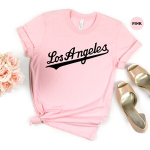 May include: A pink t-shirt with the text "Los Angeles" in a black baseball font.