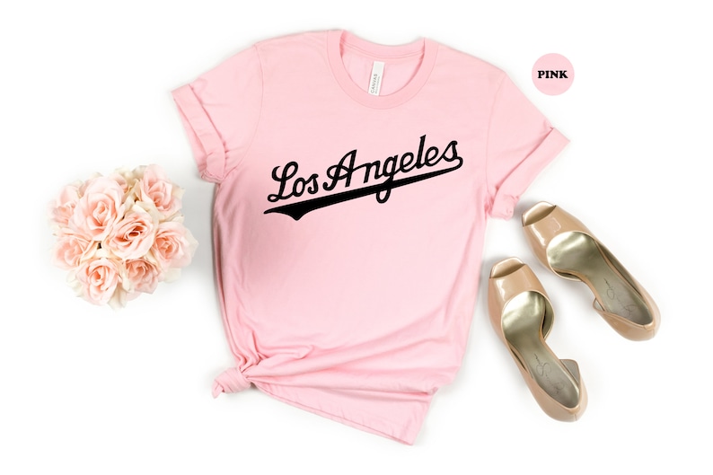 May include: A pink t-shirt with the text "Los Angeles" in a black baseball font.