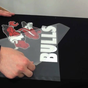 May include: Black t-shirt with a red and white graphic of a bull and the word "BULLS" in white. A person is applying a heat transfer to the shirt. The shirt is on a heat press.