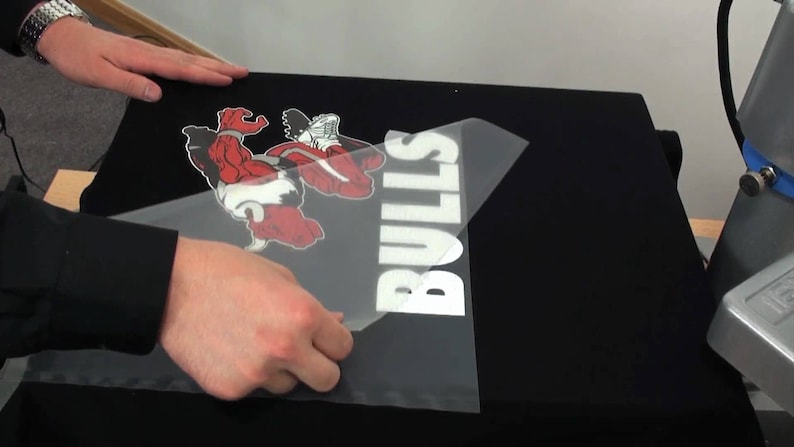 May include: Black t-shirt with a red and white graphic of a bull and the word "BULLS" in white. A person is applying a heat transfer to the shirt. The shirt is on a heat press.