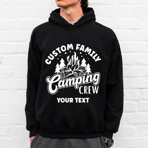 Pode incluir: Camisola de capuz preta com texto branco que diz "Custom Family Camping Crew" e "Your Text". O design apresenta uma fogueira com chamas e &aacute;rvores.