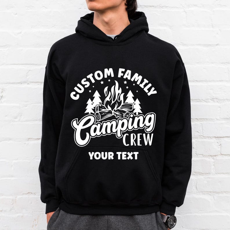 Pode incluir: Camisola de capuz preta com texto branco que diz "Custom Family Camping Crew" e "Your Text". O design apresenta uma fogueira com chamas e &aacute;rvores.