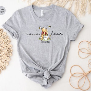 Pu&ograve; includere: T-shirt grigio chiaro con una grafica di Winnie the Pooh che abbraccia un vasetto di miele, circondato da api. Il testo recita "mama bear" sopra e "EST. 2023" sotto. La maglietta &egrave; annodata in vita.