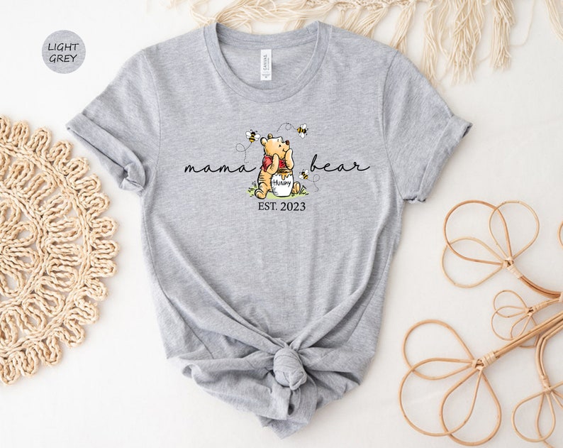 Pu&ograve; includere: T-shirt grigio chiaro con una grafica di Winnie the Pooh che abbraccia un vasetto di miele, circondato da api. Il testo recita "mama bear" sopra e "EST. 2023" sotto. La maglietta &egrave; annodata in vita.