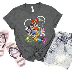 May include: A dark gray t-shirt featuring a colorful graphic of Disney characters, including Mickey and Minnie Mouse, Donald Duck, and Daisy Duck. The word "Disney" is written below. The image also includes denim shorts and striped flip-flops.