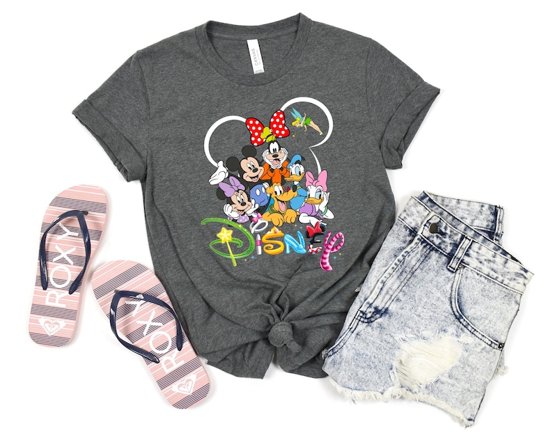 May include: A dark gray t-shirt featuring a colorful graphic of Disney characters, including Mickey and Minnie Mouse, Donald Duck, and Daisy Duck. The word "Disney" is written below. The image also includes denim shorts and striped flip-flops.