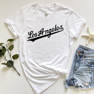May include: White t-shirt with black text that says "Los Angeles" in a baseball-style font.