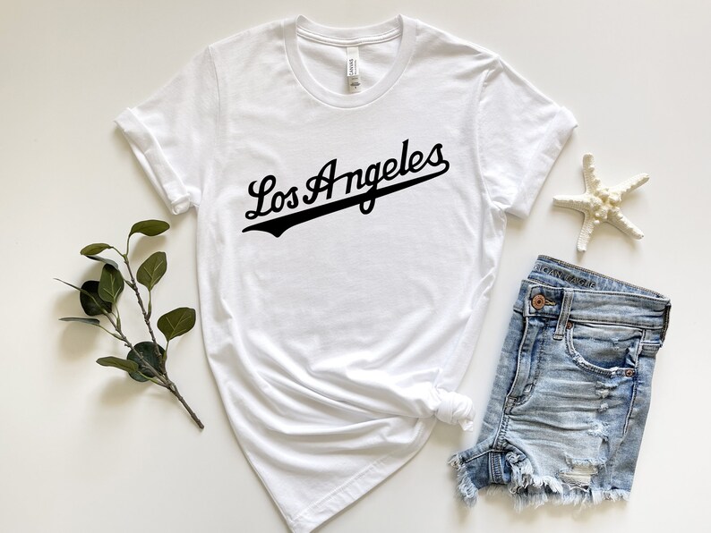 May include: White t-shirt with black text that says "Los Angeles" in a baseball-style font.