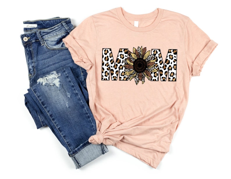 May include: A peach-colored t-shirt with the word "MOM" in black letters. The letters are decorated with a leopard print and a sunflower graphic.