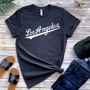May include: A dark gray t-shirt with white text that says "Los Angeles" in a baseball-style font. The shirt is laid out on a wooden surface with a pair of black sandals, a black baseball cap, and blue jeans.