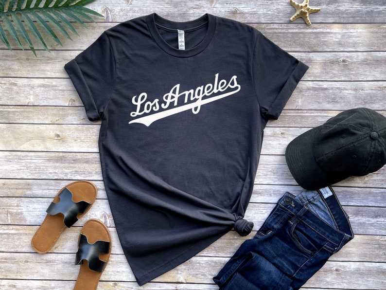 May include: A dark gray t-shirt with white text that says "Los Angeles" in a baseball-style font. The shirt is laid out on a wooden surface with a pair of black sandals, a black baseball cap, and blue jeans.