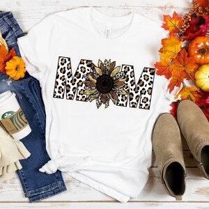 May include: White t-shirt with the word "MOM" in black letters with a leopard print pattern. A sunflower with brown and yellow petals is in the center of the word.